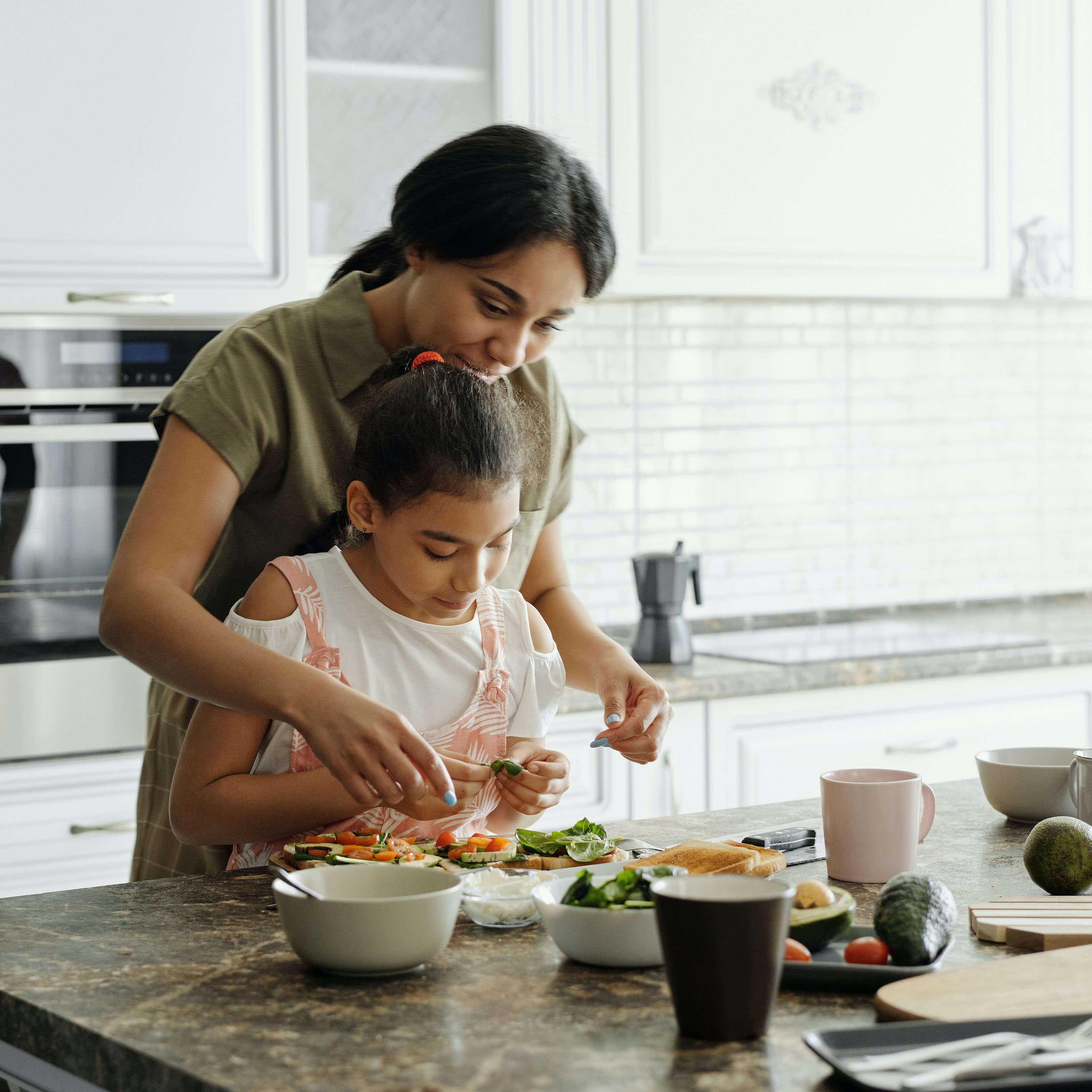 A mother standing behind her daughter and helping her prepare dinner.